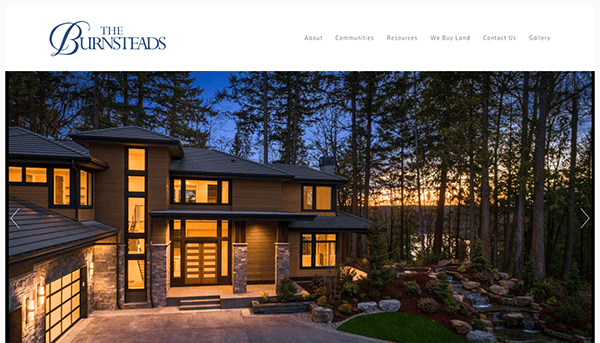 A modern, multi-story house with large windows, located in a wooded area at dusk, featured on The Burnsteads website.