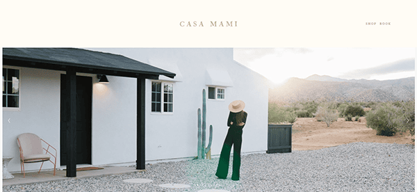 Person in black outfit and hat stands in front of a white house with black trim, next to a cactus, in a desert landscape. The sky is cloudy, and mountains are visible in the distance.