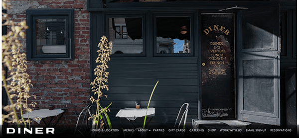 Exterior of a rustic diner with a brick wall and green door. Two small tables with chairs are placed outside. Signs list various meal options including dinner, lunch, and brunch.