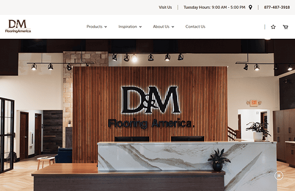 A modern reception area featuring a large, backlit "DM Flooring America" sign on a wood-paneled wall behind a marble reception desk. The space is well-lit with overhead track lighting.