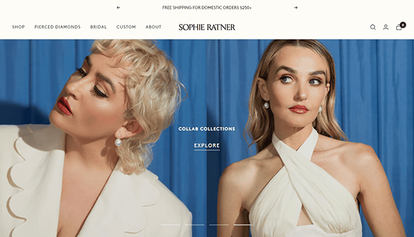 Two models in elegant attire pose against a blue backdrop, showcasing jewelry on the Sophie Ratner website. The left model wears a white blazer, and the right model wears a white halter dress.