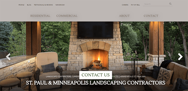 Outdoor living area with a stone fireplace, surrounded by comfortable seating and greenery. Text on image: "St. Paul & Minneapolis Landscaping Contractors." Button reads: "Contact Us.