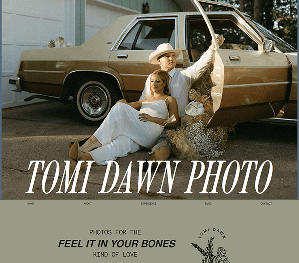 A couple poses by a beige vintage car in front of a garage. The woman sits on the ground leaning against the car, while the man stands holding the car door. Text overlay reads "Tomi Dawn Photo.