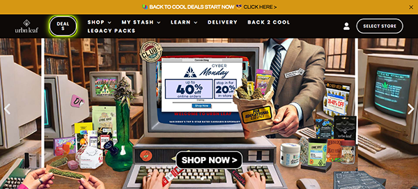 A cluttered desk with vintage computers displays a "Cyber Monday" sale ad. The screen reads, "40% off" and "Indica Happy Hour 20% off." Various cannabis products and paraphernalia are scattered around.