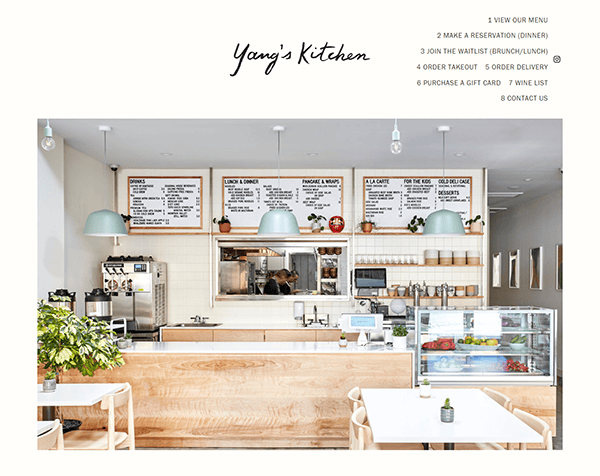 Clean and modern restaurant interior with light wood furniture and white walls. A menu board is displayed above the counter, and there is a glass case with desserts. The sign reads “Yang’s Kitchen.”.