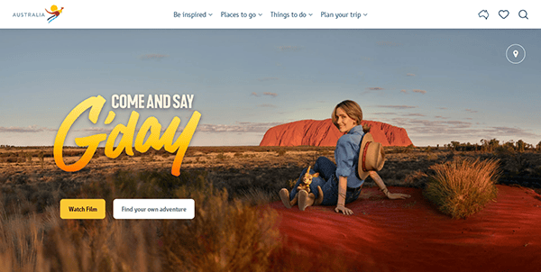 A person sitting on a red dirt landscape with a large rock formation in the background, accompanied by the text "Come and Say G'day." Website options like "Be Inspired" and "Plan Your Trip" are visible.