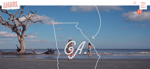 Two people walk along a beach with driftwood. The outline of Georgia state with "GA" is superimposed in the center. The text "Explore Georgia" appears at the top left.