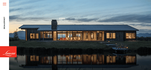 A modern single-story house with large windows and a metal roof is situated by a reflective body of water at dusk. The house has an illuminated interior and a stone chimney. Mountains are visible in the background.