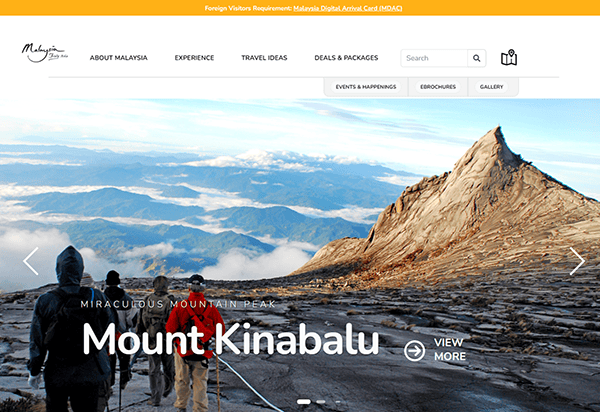 A group of hikers walks up a rocky trail towards the peak of Mount Kinabalu under a clear blue sky. Text on the image reads "Mount Kinabalu" and site navigation links are visible at the top.