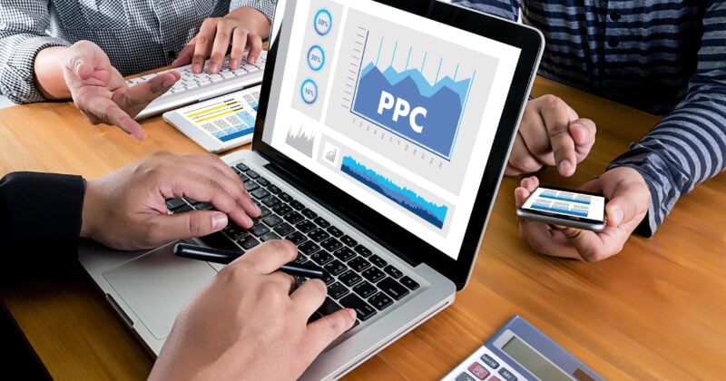 People working at a desk with a laptop displaying a PPC graph, holding smartphones and using a calculator, focus on PPC essentials to maximize leads for plumbing companies.