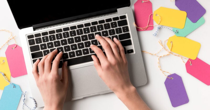 Hands typing on a laptop keyboard, surrounded by colorful blank tags on strings, each tag cleverly illustrating Google Display Ads Sizes for maximum impact.