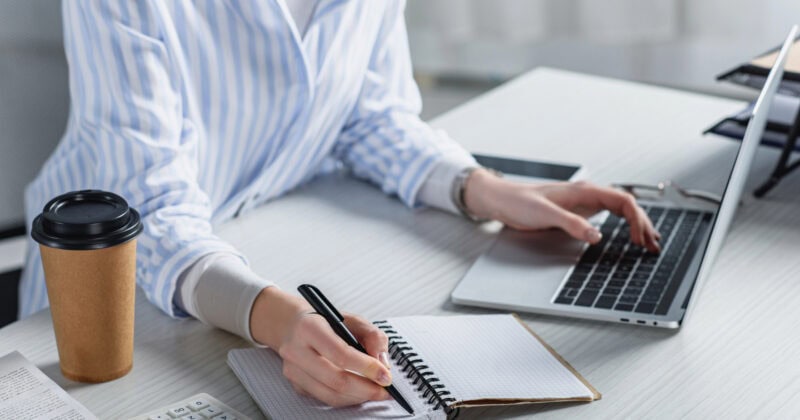 Cropped view of woman writing in notebook and holding laptop