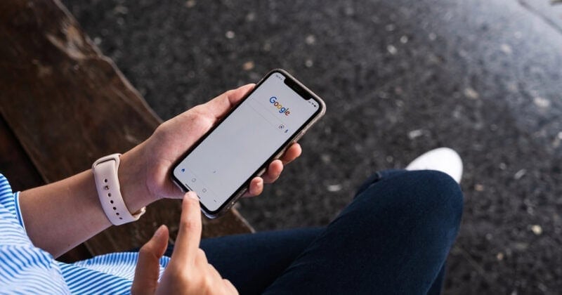 A person sits on a bench, dark pavement beneath them, holding a smartphone with the Google search page open. It's the perfect moment to craft high-conversion Google Ads or find that ideal copy template for digital campaigns.