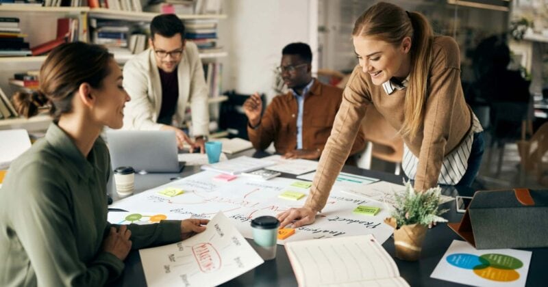 Four people collaborating at a table with charts, documents, and a laptop in a modern office setting, discussing advanced remarketing services to enhance their retargeting tactics.