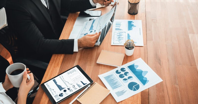 Expert marketers in business attire are reviewing charts and graphs at a wooden table, strategically planning their retargeting campaigns over a tablet, with notebooks and a cup of coffee by their side.