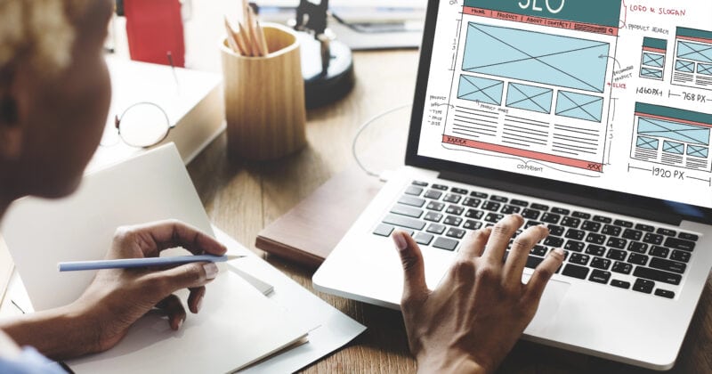 A person is working on a laptop displaying an SEO webpage layout, surrounded by notebooks and pencils, while exploring advanced PPC Reporting Tools.