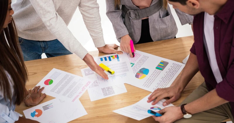 A group of people collaborates at a table, highlighting charts and documents with markers as they develop an advanced strategy for remarketing campaigns.