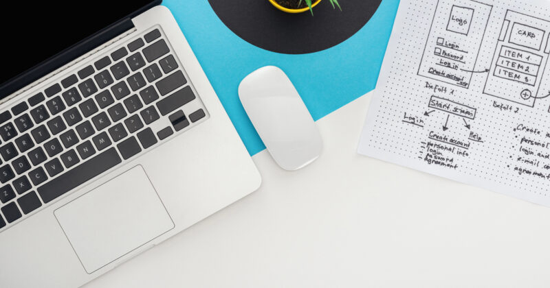 A laptop, wireless mouse, a small potted plant, and a sheet of paper with B2B website design ideas are neatly arranged on the white and blue desk.