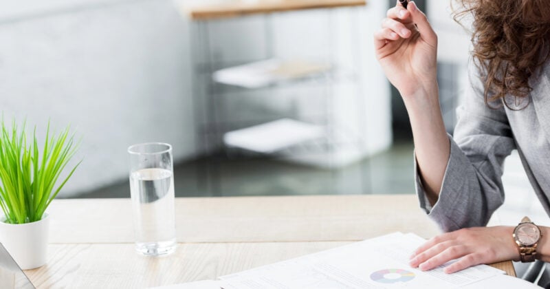 A person holds a pen, reviewing documents with a game-changing colorful chart at a desk. Nearby are a glass of water, a small green plant, and part of a laptop, tools every professional developer values to stay on top in the world of web development.