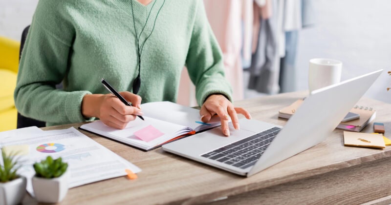 A person in a green sweater, focused on marketing strategies, takes notes at a desk while working on a laptop, surrounded by documents and plants.