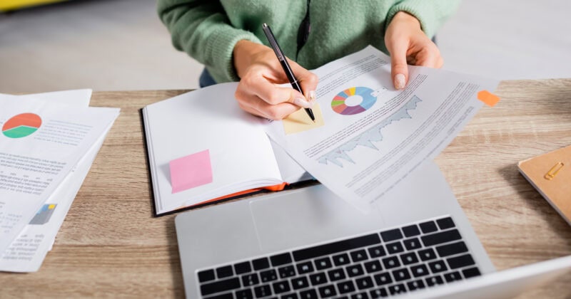 Person analyzing charts at a desk with a laptop, notebook, and documents, focused on aligning advertising solutions to strategic goals.