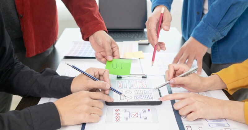 A group of hands collaboratively refining a web design plan, with sketches and colored notes detailing best practices and user experience insights spread across the table.