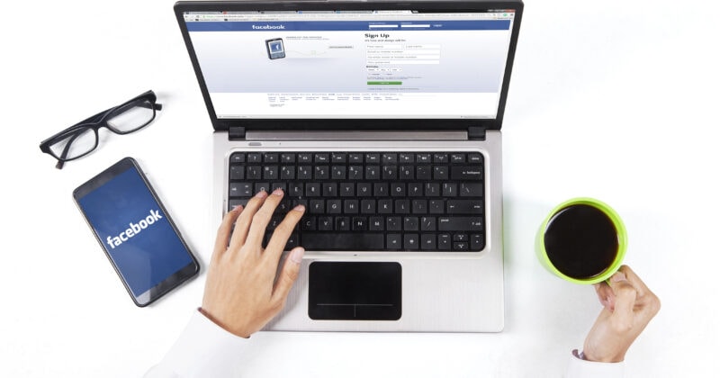 A person uses a laptop with the Facebook sign-up page open, while their smartphone showcases the Facebook logo. Nearby are glasses and a coffee cup—ideal for pondering proven strategies to enhance campaign performance through precise Facebook audience targeting.