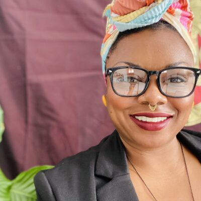 A smiling person wearing glasses and a colorful headwrap, with a septum ring and a necklace shaped like a map. A plant is visible in the background.