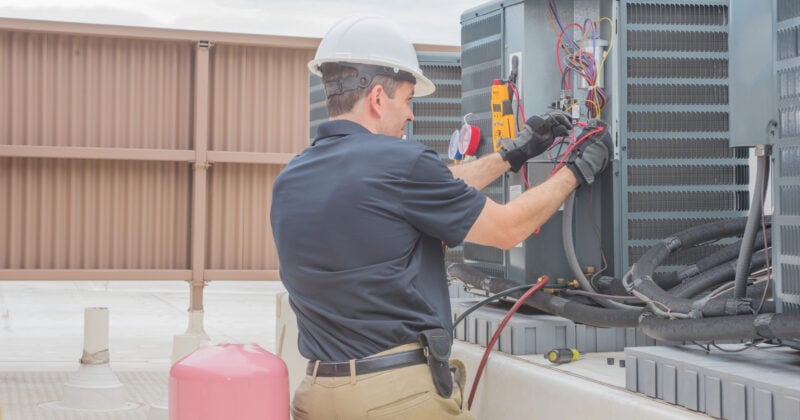 A technician from leading HVAC companies, wearing a helmet and gloves, kneels to repair an HVAC unit on a rooftop, surrounded by tools and equipment.