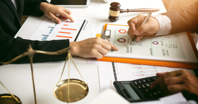 Two people reviewing financial charts and graphs on a clipboard, with a gavel, scales, calculator, and coffee cup on the table—ready for any accounting firm website looking to showcase professionalism.