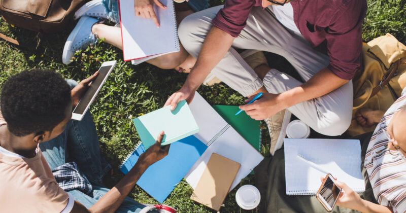 A group of people sitting on grass, holding notebooks, tablets displaying college websites, and coffee cups, engaged in discussion.