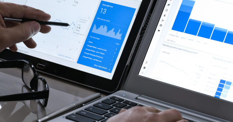 Two people analyze graphs on tablet and laptop screens, one pointing with a pen, optimizing SEO keywords for service businesses. Glasses, smartphone, and pen lie nearby on a table.