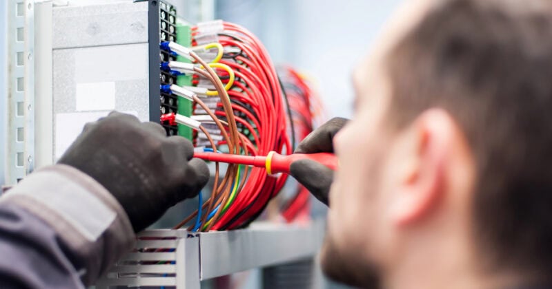 Person wearing gloves uses a screwdriver to work on a panel with numerous red and colored wires.