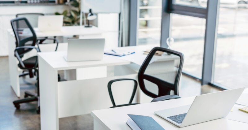 Sunlight streams through large windows into a modern office with white desks, black chairs, laptops, and a notebook. Perfect for companies who've used proven strategies to optimize space, creating an inspiring and efficient work environment.