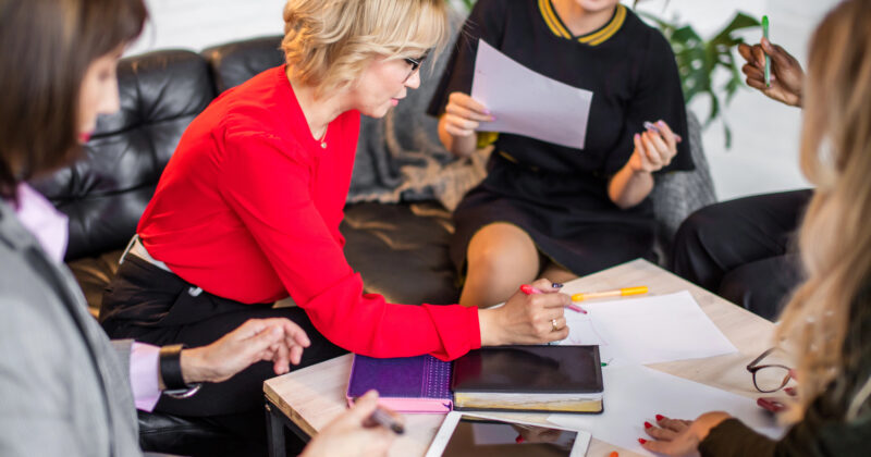 Five entrepreneurs collaborate at a table with papers, pens, and digital tablets. A female in a red top focuses on writing, while others discuss and exchange documents, exemplifying the dynamic spirit of today's female entrepreneurs.