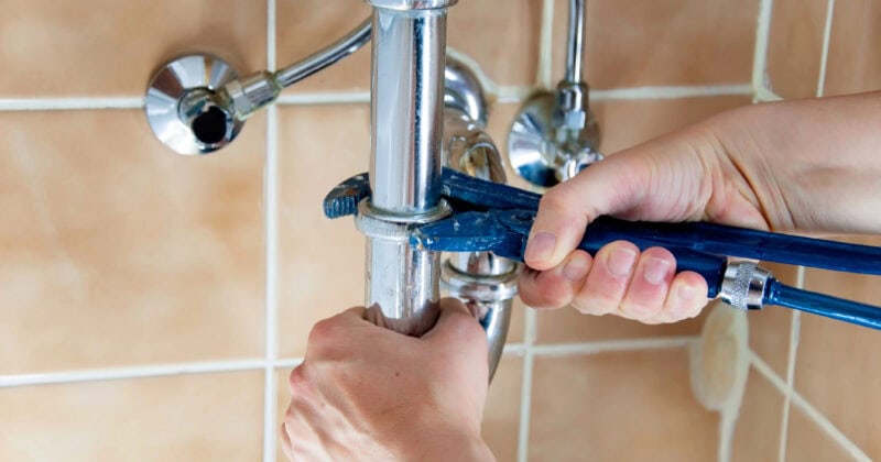 A person expertly uses a blue wrench to tighten a pipe beneath a sink, showcasing essential techniques against a tiled wall background.