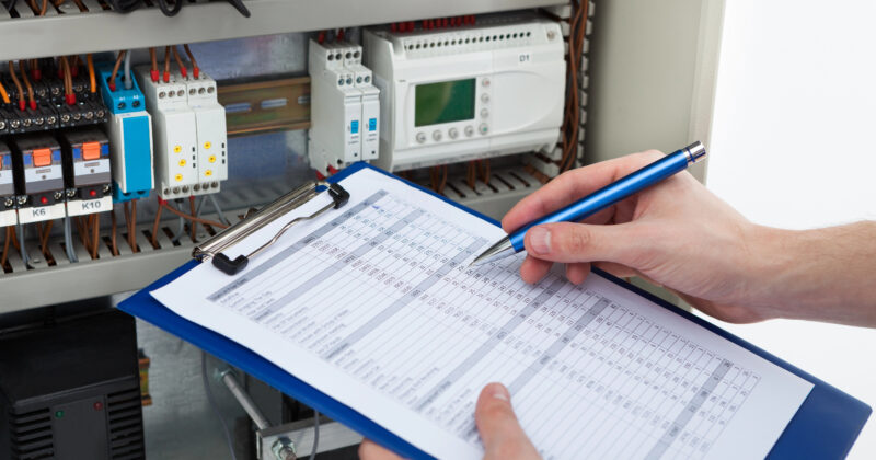 An electrician with a blue pen and clipboard is recording data in the service area. Nearby, an open electrical panel reveals a maze of wires and various components.