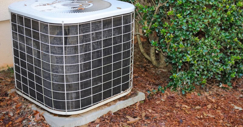 The outdoor air conditioning unit, nestled beside the building, is surrounded by mulch and lush greenery. Its strategic placement ensures efficiency and blends seamlessly with the landscape, making it a key element in any construction strategy that aims to convert visitors into admirers.