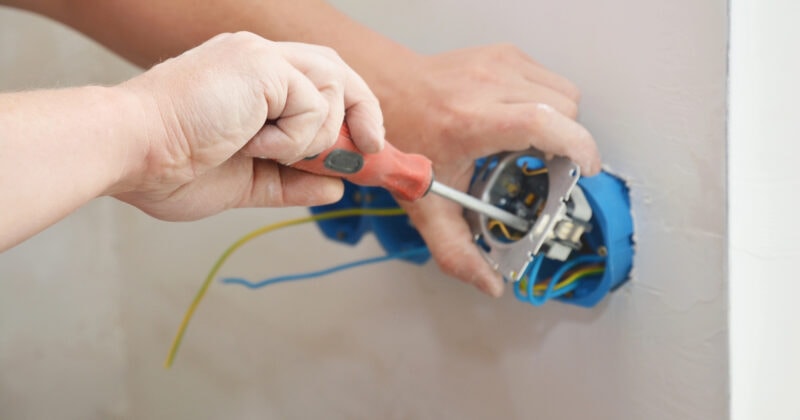 A person skillfully uses a screwdriver to repair the electrical wiring in a wall socket, perfectly embodying the precision and expertise that one might feature on an Electrician Website Design.