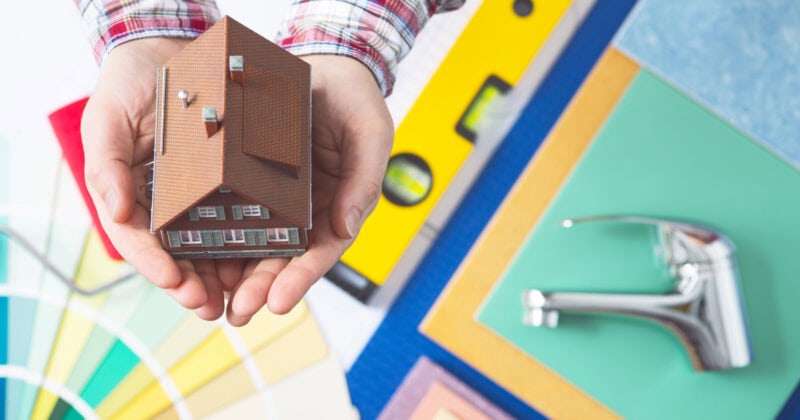 Hands holding a small model house, surrounded by a color palette, a faucet, and a level, suggesting home renovation or real estate planning to maximize ROI.