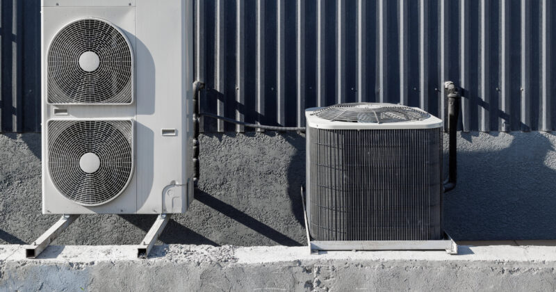 Two outdoor HVAC units rest on a concrete slab against a corrugated metal wall, one unit vertically oriented and the other horizontally.