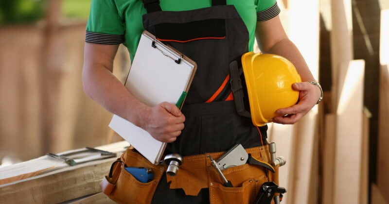 A person in a green shirt and black overalls holds a clipboard and yellow hard hat, equipped with a tool belt filled with various tools—an emblem of effective website design principles for 2024, echoing the precision found in home builder website design.