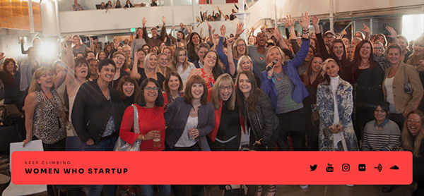 A large group of diverse female entrepreneurs is smiling and raising their hands at a Women Who Startup event, with a speaker holding a microphone in the middle.