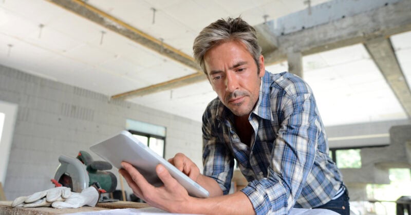 A man in a plaid shirt leans on a wooden surface, engrossed in his tablet as he reviews blueprints. The room under construction buzzes with potential, reflecting the meticulous planning home builders bring to life for their clients.