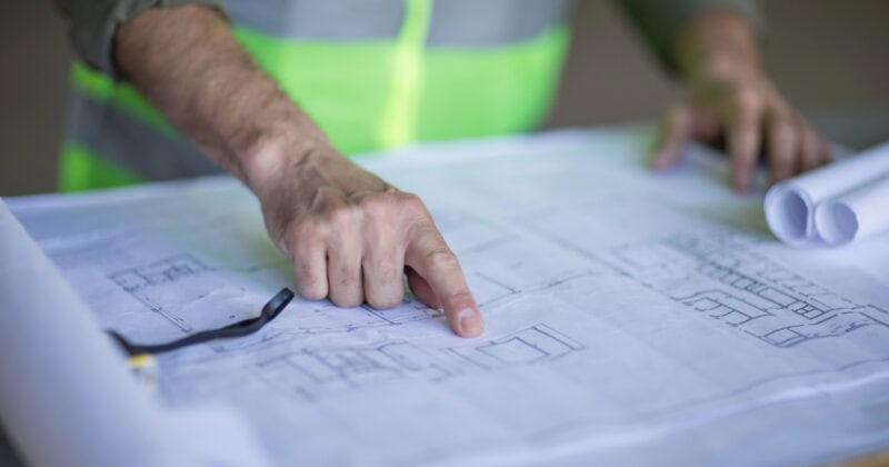 A person in a reflective vest points at architectural blueprints on a table, highlighting the innovative design strategies for an upcoming contractor marketing project.