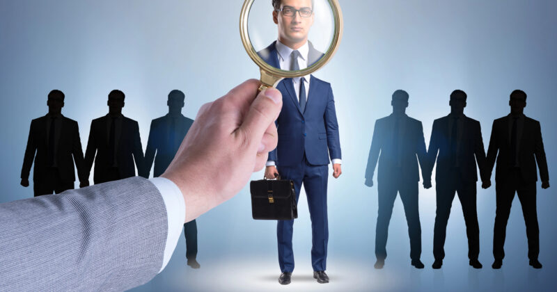 A hand holding a magnifying glass focuses on a man in a suit, standing among silhouettes of similar figures. With a briefcase in hand, he suggests a business context, much like using the best background check to discern standout professionals from the crowd.