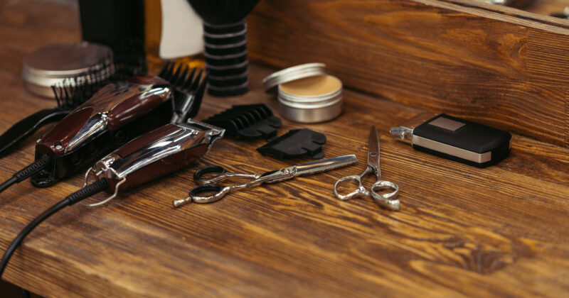 Barber tools on a wooden counter, including electric clippers, scissors, a comb, a brush, and the trimmer used by city's best barber shop. A mirror reflects some of the items.