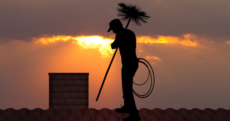 Silhouette of a chimney cleaner at sunset, holding a brush and a coil of rope, standing on a roof.