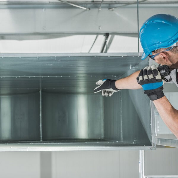 A worker in a blue helmet inspects a large metal ventilation duct, pointing and shining a flashlight inside, standing on a ladder in an industrial setting.