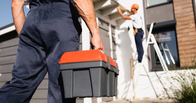 A worker in uniform holds a toolbox in the foreground while another person in uniform paints a house using a ladder in the background, exemplifying the commitment to quality that defines a trusted home builder.